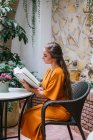 Вид сбоку нежной женщины, читающей интересную книгу, сидя за столом на летней террасе дома — стоковое фото
