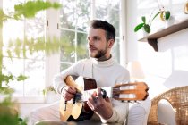 Besinnlicher männlicher Musiker mit Tätowierungen, der klassische Gitarre spielt, während er im Sessel sitzt und gegen das Fenster im Haus blickt — Stockfoto