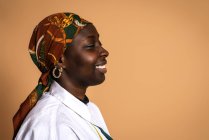 Modèle féminin afro-américain joyeux en foulard tendance et chemise blanche riant avec les yeux fermés sur fond beige en studio — Photo de stock
