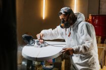 Männliche Shaper in Atemschutzmaske und Kostüm mit elektrischem Hobel und Polierfläche des Surfbretts in der Werkstatt — Stockfoto