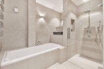 Diseño interior contemporáneo de baño ligero con bañera blanca y amplia cabina de ducha decorada con azulejos grises con iluminación - foto de stock