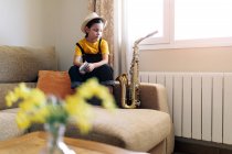 Удумлива дитина в капелюсі текстових повідомлень на мобільному телефоні, сидячи на дивані з саксофоном у вітальні, дивлячись далеко — стокове фото