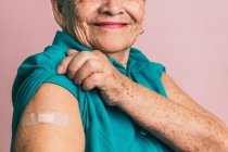 Récoltée positive femme âgée méconnaissable montrant bras avec patch après la vaccination de COVID sur fond rose et regardant la caméra — Photo de stock