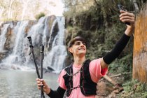 Caminhante do sexo masculino sorridente tomando auto-tiro no smartphone enquanto estava no fundo da cachoeira e lago na floresta durante o trekking — Fotografia de Stock