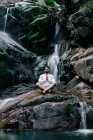 Tranquilo hombre sentado en pose de loto con las manos de oración en la roca cerca de la cascada y meditando mientras hace yoga con los ojos cerrados - foto de stock