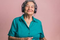 Alegre mujer mayor moderna escuchando música en auriculares y bailando con los ojos cerrados sobre fondo rosa en el estudio - foto de stock