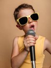 Cool child in sunglasses singing in modern mic on brown background in studio — Stock Photo
