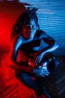 Sonhador preto músico masculino com torso nu tocando tambor africano em estúdio com luzes de néon vermelho e azul — Fotografia de Stock