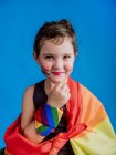 Menina sorridente com bochecha pintada com bandeira multicolorida em fundo azul vívido — Fotografia de Stock