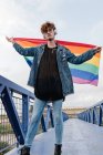 Maschio omosessuale elegante con bandiera LGBT colorata in piedi sul ponte e ascoltare musica in cuffia mentre guarda la fotocamera — Foto stock