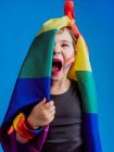 Menina bonito com lábios vermelhos e bandeira do arco-íris cobrindo metade da cabeça olhando para a câmera no fundo azul enquanto ela grita — Fotografia de Stock