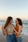 Young girlfriends embracing each other while standing on sandy beach near waving sea at sundown looking at each other — Stock Photo