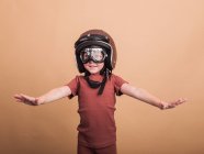 Веселый ребенок в шлеме и защитных очках смотрит в камеру с протянутыми руками на бежевом фоне — стоковое фото
