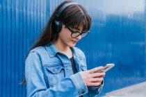 Позитивна жінка в навушниках, які переглядають смартфон, стоячи на синьому фоні на вулиці — стокове фото