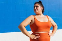 Избыточный вес этническая спортсменка в активной износа глядя в сторону синей плитки стены в солнечный день — стоковое фото