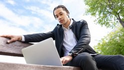 De dessous de freelance masculin ethnique avec coiffure tressée assis sur le banc et tapant sur le clavier de l'ordinateur portable tout en travaillant sur le projet à distance sur la rue urbaine en utilisant des écouteurs TWS — Photo de stock