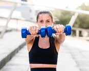 Soft focus of determined female athlete doing exercise with dumbbells during fitness workout on city street in summer — Stock Photo
