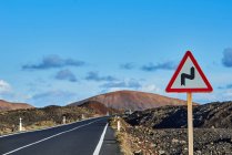 Дорожный знак, предупреждающий о двойном изгибе дороги, расположенный рядом с асфальтовым шоссе на фоне дождей и облачного голубого неба в Фуэртевентуре, Испания — стоковое фото
