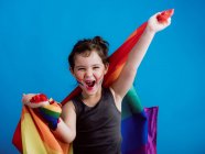 Усміхнена дівчина з розмальованою щокою піднімає руки з різнокольоровим прапором на яскраво-блакитному фоні — стокове фото