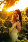 Side view of graceful young Hispanic female in stylish yellow dress standing amidst blooming sunflowers in countryside field in sunny summer day looking at camera — Stock Photo