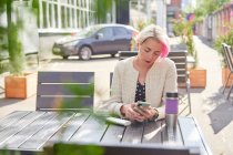 Alternative féminine aux cheveux courts naviguant sur les médias sociaux sur smartphone alors qu'elle était assise à table dans un café de rue le jour ensoleillé — Photo de stock