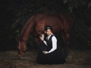 Fiduciosa signora afroamericana adulta in abiti eleganti e cappello seduto con cavallo marrone, mentre guardando lontano vicino agli alberi di giorno — Foto stock