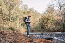 Caminhante masculino com mochila em pé em solo rochoso perto de cachoeira na floresta e olhando para longe — Fotografia de Stock