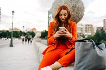 Mujer elegante feliz con el pelo rojo y en traje naranja vibrante sentado en la frontera de piedra en la ciudad y mensajería en el teléfono celular - foto de stock