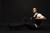 Vista lateral do jovem modelo masculino hipster étnico confiante com cabelo trançado vestindo camisa branca com roupas pretas olhando para a câmera enquanto sentado no estúdio escuro — Fotografia de Stock