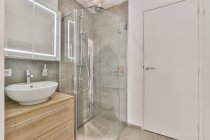 Стильний білий інтер'єр ванної кімнати з умивальником і дерев'яним шафою біля душової кабіни зі скляною перегородкою в сучасній квартирі — стокове фото