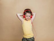 Niño preadolescente despreocupado en auriculares rojos escuchando música mientras está de pie sobre un fondo marrón y mirando a la cámara - foto de stock