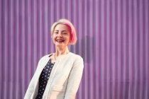 Charming alternative female with dyed hair and in trendy clothes standing against violet wall in city and looking at camera — Stock Photo