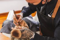 Hoher Winkel des tätowierten Mannes in Maske zeichnet Eyeliner-Pfeile auf den Augenlidern der Frau bei der Arbeit im Make-up-Studio — Stockfoto
