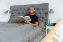 Maschio pensieroso seduto sul letto morbido al mattino e leggere storia interessante nel libro dopo il risveglio — Foto stock