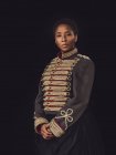 Femme adulte afro-américaine confiante dans une veste élégante regardant la caméra dans un studio sombre sur fond noir — Photo de stock