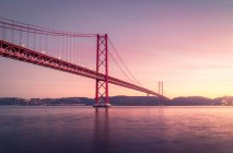 Famoso puente 25 de Abril que cruza el río Tajo y conecta Lisboa y Almada cerca del Santuario de Cristo Rey monumento contra el cielo nublado al atardecer en Portugal - foto de stock