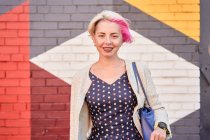 Happy alternative female in trendy dress and with dyed short hair standing against colorful brick wall in street and looking at camera — Stock Photo