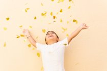 Счастливый мальчик в белой футболке стоит с поднятыми руками, бросая вверх кучу желтых услышать конфетти на светло-оранжевой стене — стоковое фото