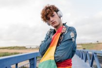 Homosexual elegante hombre con colorida bandera LGBT de pie en el puente y escuchar música en los auriculares mientras mira a la cámara - foto de stock