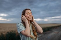 Стильная счастливая молодая женщина с закрытыми глазами, касающаяся длинных волос на проезжей части под облачным небом в сумерках — стоковое фото