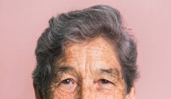 Meia face cortada de mulher idosa com cabelos grisalhos curtos e olhos castanhos olhando para a câmera em fundo rosa em estúdio — Fotografia de Stock