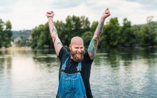 Happy bald male with beard and tattoos in casual clothes standing with raised arms near lake and green trees in summer — Stock Photo