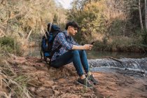 Vista laterale del contenuto escursionista maschio con zaino seduto vicino al lago nei boschi e la navigazione cellulare durante il viaggio — Foto stock