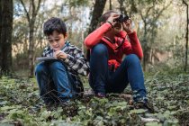Ethnic boy writing in notepad against sister looking through binoculars while sitting on land in summer woods — Stock Photo