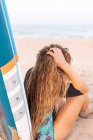 Back view of unrecognizable female surfer sitting with blue SUP board on sandy seashore in summer and looking away — Stock Photo