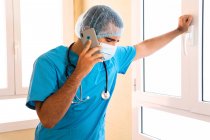 Мужчина-медик в форме и со стетоскопом, стоящим в больнице и разговаривающим по смартфону — стоковое фото