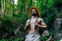 D'en bas du voyageur homme torse nu avec sac à dos et chapeau debout dans les bois rocheux et regardant loin — Photo de stock
