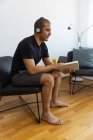 Продуманий чоловік читає цікаву книгу, сидячи на стільці у вітальні вранці вдома з навушниками — стокове фото