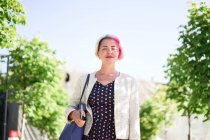 Mulher alternativa encantada com cabelo tingido em pé na rua no dia ensolarado no verão e olhando para a câmera — Fotografia de Stock