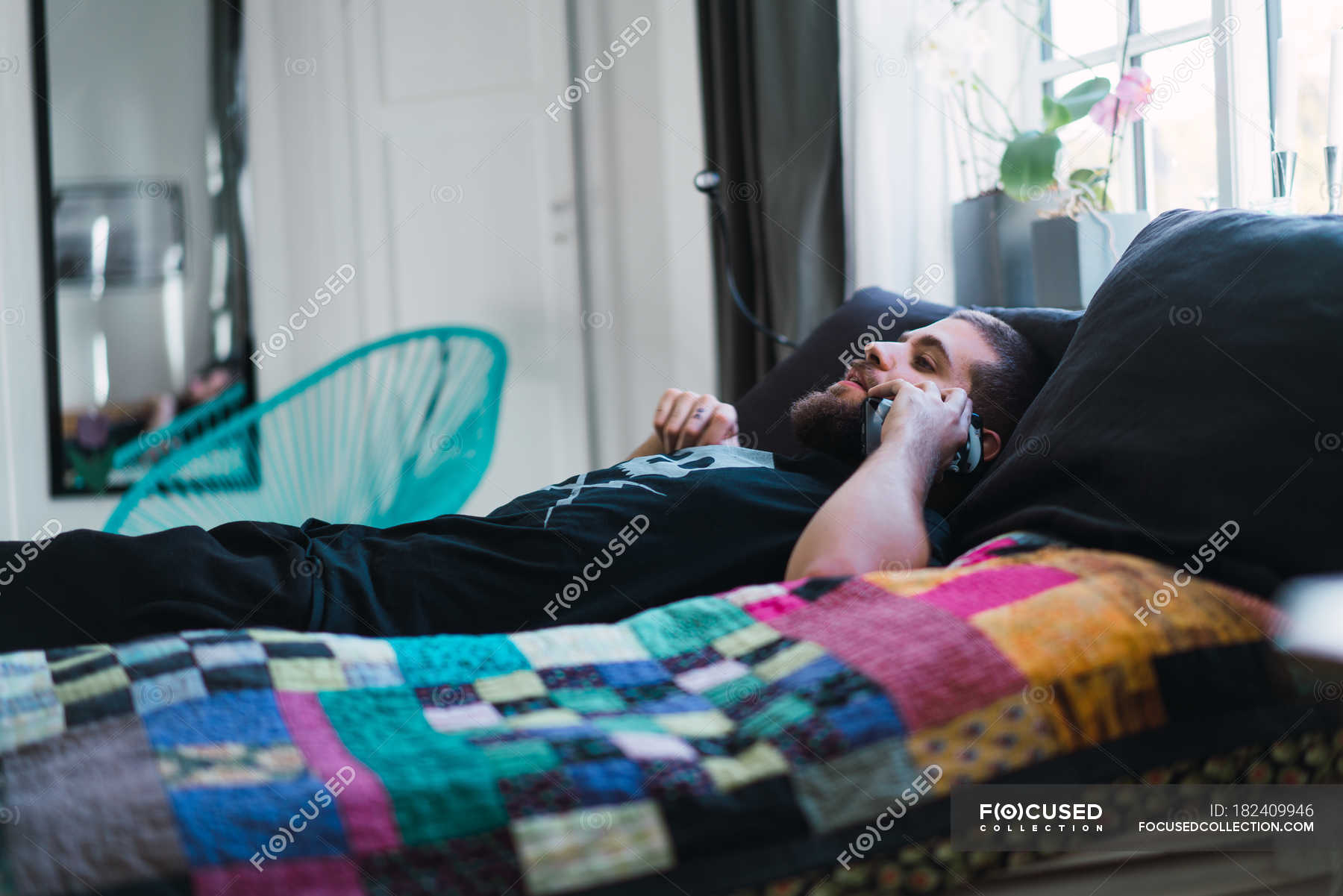 Side view of man lying on bed and talking on smartphone at home ...